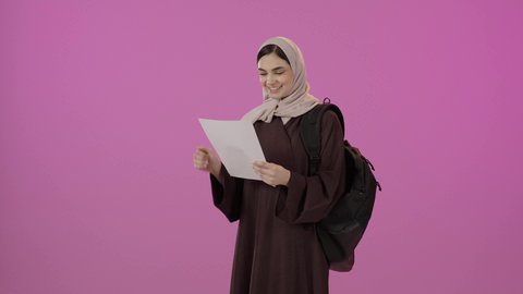 Gestures of happiness and joy, higher education in universities and academic colleges, preparation for the new academic year, a portrait of a Gulf Arab Saudi female university student wearing a hijab and an abaya, holding papers in one hand and raising her fist with expressions of victory and achieving goals, colorful background.