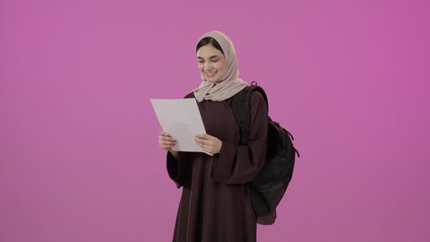 Preparation for the new academic year, higher education in universities and academic colleges, gestures of happiness and joy, a portrait of a Gulf Arab Saudi university student wearing a hijab, dressed in an abaya and carrying a backpack, holding papers in one hand and raising a fist with expressions of victory and achieving goals, colorful background.