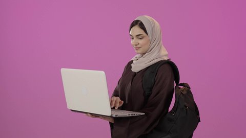 مفهوم التعليم عن بعد، استخدام الأجهزة التقنية الحديثة لمتابعة المحاضرات، بورتريه لطالبة جامعية عربية خليجية سعودية محجبة تحمل حقيبة الظهر والحاسوب المحمول، خلفية ملونه