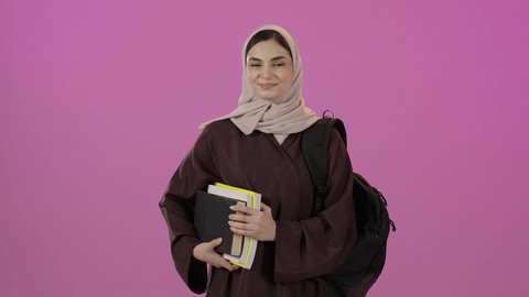 The feeling of pride and self-confidence, the concept of diligence and excellence, expressions of acceptance and approval, a Gulf Arab Saudi student wearing an abaya and carrying a bag with a collection of books, looking at the camera with gestures of happiness and joy, a colorful background.