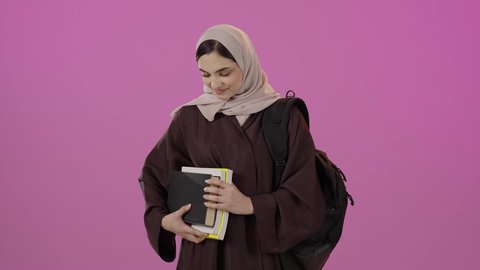 مفهوم الاجتهاد والتفوق، الشعور بالفخر والثقة بالنفس، تعابير القبول والموافقة، طالبة عربية خليجية سعودية ترتدي عباءه وحقيبة تحمل مجموعه من الكتب ، النظر الى الكاميرا بايماءات السعادة و السرور، خلفية ملونه