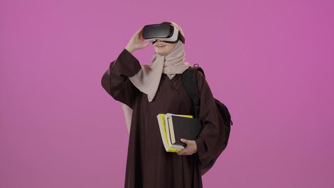 Gestures of happiness and joy, education using virtual reality technology, simulating the imaginary world, integrating modern technologies into learning and training, a portrait of a university student, an Arab Gulf Saudi woman wearing a hijab, carrying a backpack, wearing 3D virtual reality glasses, holding a collection of books, raising her hand to try to touch something, colorful background.