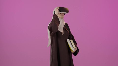 التعليم باستخدام تقنية العالم الافتراضي، محاكاة العالم الخيالي، دمج التقنيات الحديثة بالتعلم والتدريب، بورتريه لطالبة جامعية عربية خليجية سعودية محجبة ترتدي حقيبة الظهر تضع نظارة الفي ار ثلاثية الأبعاد تحمل مجموعة من الكتب ترفع يدها لمحاولة لمس شيء ما، ايماءات السعادة و السرور ، خلفية ملونه