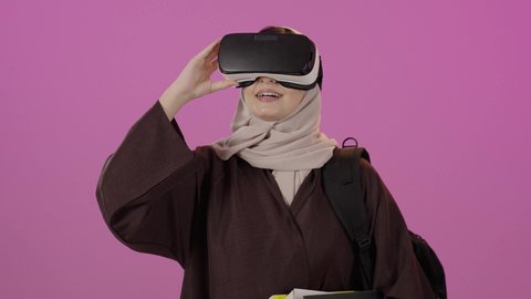 Integrating modern technologies with learning and training, simulating the imaginary world, education using virtual reality technology, a portrait of a university student, an Arab Gulf Saudi woman wearing a hijab, carrying a backpack, wearing 3D VR glasses, holding a collection of books, raising her hand to try to touch something, gestures of happiness and joy, colorful background.