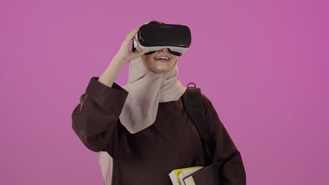 محاكاة العالم الخيالي، دمج التقنيات الحديثة بالتعلم والتدريب، التعليم باستخدام تقنية العالم الافتراضي، بورتريه لطالبة جامعية عربية خليجية سعودية محجبة ترتدي حقيبة الظهر تضع نظارة الفي ار ثلاثية الأبعاد تحمل مجموعة من الكتب ترفع يدها لمحاولة لمس شيء ما، ايماءات السعادة و السرور ، خلفية ملونه