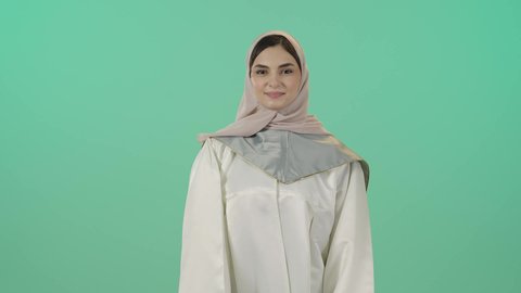 الوقوف بثقه بأيدي متقاطعه، بورتريه لطالبة عربية سعودية خليجية ترتدي العباءه وقبعة التخرج, ايماءات تدل على الشعور بالفخر والسعاده,  مفهوم التفوق والنجاح, التعليم في السعوديه, خلفية خضراء