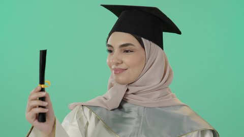 حفل التخرج الجامعي، النظر الى الكاميرا بايماءات البهجة والسعادة بالوصول لحلم النجاح، بورتريه مقرب لخريجة عربية خليجية سعودية محجبة مبتسمة ترتدي العباءة وقبعة التخرج تمسك شهادة التفوق وترفع قبضة اليد بتعابير الفوز وتحقيق الاهداف، خلفية خضراء