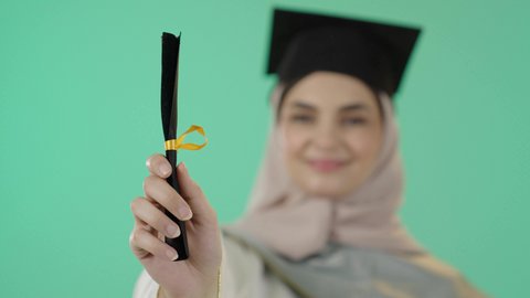 المناسبات والاحتفالات الجامعية، النظر الى الكاميرا بايماءات البهجة والسعادة بالوصول لحلم النجاح، بورتريه مقرب ليد خريجة عربية خليجية سعودية محجبة مبتسمة ترتدي العباءة وقبعة التخرج تمسك شهادة التفوق، خلفية خضراء