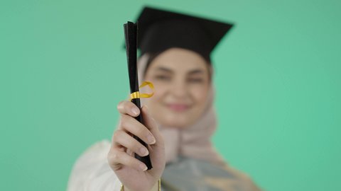 الاحتفال بتحقيق النجاح، النظر الى الكاميرا بايماءات البهجة والسعادة بالوصول لحلم التخرج، بورتريه مقرب ليد خريجة عربية خليجية سعودية محجبة مبتسمة ترتدي العباءة وقبعة التخرج تمسك شهادة التفوق، خلفية خضراء