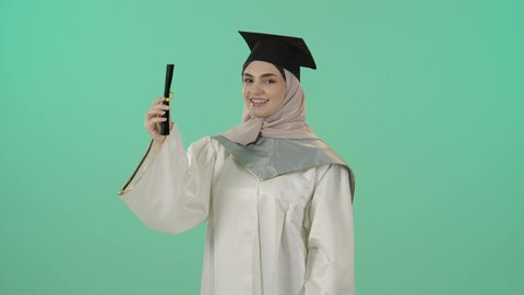 النظر الى الكاميرا بايماءات البهجة والسعادة بالوصول لحلم النجاح، بورتريه لخريجة عربية خليجية سعودية محجبة مبتسمة ترتدي العباءة البيضاء وقبعة التخرج تمسك بيدها شهادة التفوق وترفع قبضة اليد بتعابير الفوز وتحقيق الاهداف، خلفية خضراء