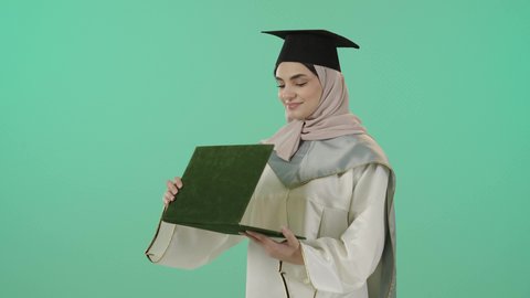 الاحتفال بتحقيق النجاح, أهمية الدراسة الجامعية, ايماءات البهجة والسعادة بالوصول لحلم التخرج، بورتريه لخريجة عربية سعودية خليجية محجبة ترتدي العباءة البيضاء وقبعة التخرج تمسك بيدها شهادة التفوق و تنظر اليها بتعابير الفخر و الدهشه, خلفية خضراء