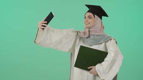 ايماءات السعادة و السرور، المناسبات والاحتفالات الجامعية، توثيق اللحظات السعيدة, بورتريه لطالبة عربية سعودية خليجية محجبة ترتدي ثوب التخرج والقبعة تمسك بيدها الوثيقة والهاتف المحمول تقوم بالتقاط صورة سيلفي تذكارية, خلفية خضراء