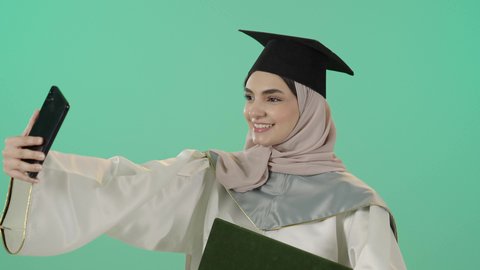 University events and celebrations, documenting happy moments, a close-up portrait of a Saudi Arabian Gulf hijabi student wearing a graduation gown and cap, holding a diploma and a mobile phone while taking a commemorative selfie, with a green background.