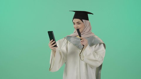 Celebrating the completion of university studies, sharing with family and friends the joy of achieving the dream of success, a portrait of a Saudi Gulf Arab graduate wearing a graduation gown and cap, holding a diploma while making a video call via a mobile phone, using modern technology, with a green background.