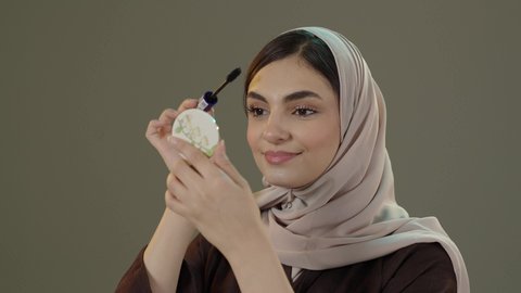 لاهتمام بجمال الشكل والهيئة، ايماءات السعادة و السرور، المستحضرات التجميل النسائية، دورات تعليمية لمكياج تتوريال، استخدام الماسكرا لإبراز جمال العينين، الإهتمام بالجمال والمظهر الخارجي، بورتريه مقرب لامراة عربية خليجية سعودية  محجبة تقوم بوضع الميكاب، خلفيه رمادية