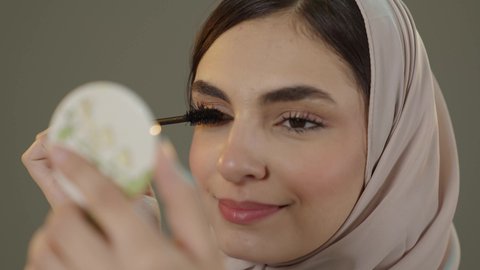 العناية بالأناقة والجاذبية الشخصية، ايماءات السعادة و السرور، المستحضرات التجميل النسائية، دورات تعليمية لمكياج تتوريال، استخدام الماسكرا لإبراز جمال العينين، الإهتمام بالجمال والمظهر الخارجي، بورتريه مقرب لامراة عربية خليجية سعودية  محجبة تقوم بوضع الميكاب، خلفيه رمادية