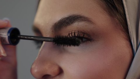 المستحضرات التجميل النسائية، دورات تعليمية لمكياج تتوريال، استخدام الماسكرا لإبراز جمال العينين، الإهتمام بالجمال والمظهر الخارجي، بورتريه مقرب لامراة عربية خليجية سعودية  محجبة تقوم بوضع الميكاب، خلفيه رمادية