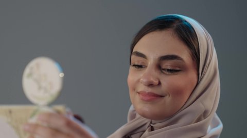 تعزيز الأنوثة بالعناية بالبشرة والجمال والإطلالة الأنيقة، استخدام المساحيق التجميلية, مفهوم التزين والتجمل, التسويق لمستحضرات التجميل, بورتريه مقرب لامراة عربية خليجية سعودية محجبة تستعمل علبة وفرشاة أحمر الخدود، خلفية رمادية