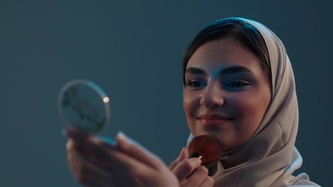 الاهتمام بالأنوثة عبر الجمال والعناية بالبشرة والإطلالة، استخدام المساحيق التجميلية, مفهوم التزين والتجمل, التسويق لمستحضرات التجميل, بورتريه مقرب لامراة عربية خليجية سعودية محجبة تستعمل علبة وفرشاة أحمر الخدود، خلفية رمادية