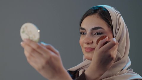إبراز الأنوثة عبر العناية الشخصية والجمال الخارجي، استخدام المساحيق التجميلية, مفهوم التزين والتجمل, التسويق لمستحضرات التجميل, بورتريه مقرب لامراة عربية خليجية سعودية محجبة تستعمل علبة وفرشاة أحمر الخدود، خلفية رمادية