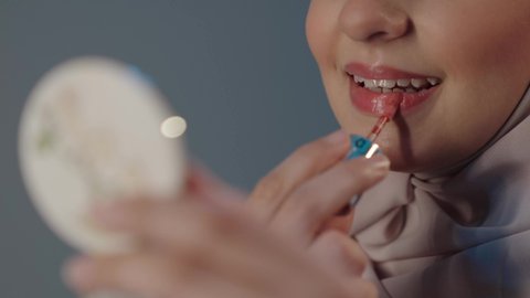 العناية بالجمال والمظهر الخارجي، الترويج لمنتجات و مساحيق التجميل، استعمال المستحضرات التجميلية، مفهوم التزين والتجمل, بورتريه مقرب لامرأة عربية خليجية سعودية محجبة ترتدي العباءة تضع أحمر شفاه, خلفية رمادية