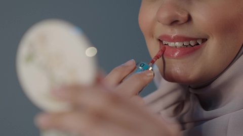 الترويج لمنتجات و مساحيق التجميل، استعمال المستحضرات التجميلية، مفهوم التزين والتجمل, العناية بالجمال والمظهر الخارجي, بورتريه مقرب لامرأة عربية خليجية سعودية محجبة ترتدي العباءة تضع أحمر شفاه, خلفية رمادية