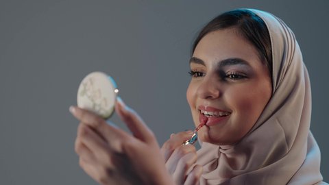 مفهوم التزين والتجمل, استعمال المستحضرات التجميلية, الترويج لمنتجات و مساحيق التجميل, العناية بالجمال والمظهر الخارجي, بورتريه مقرب لامرأة عربية خليجية سعودية محجبة ترتدي العباءة تضع أحمر شفاه, خلفية رمادية