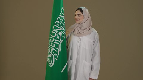 ذكرى تأسيس الدولة السعودية الأولى 22 فبراير، التباهي والتفاخر بامتلاك الهوية السعودية، اليوم الوطني السعودي، بورترية لسيدة عربية خليجية سعودية مبتسمة محجبة ترتدي العباءة تقف امام علم المملكة تنظرالية بتعابير الفخر و السرور، خلفية بني