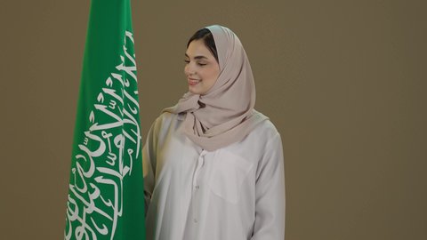 التباهي والتفاخر بامتلاك الهوية السعودية، اليوم الوطني السعودي، بورترية لسيدة عربية خليجية سعودية مبتسمة محجبة ترتدي العباءة تقف امام علم المملكة تنظرالية بتعابير الفخر و السرور، خلفية بني