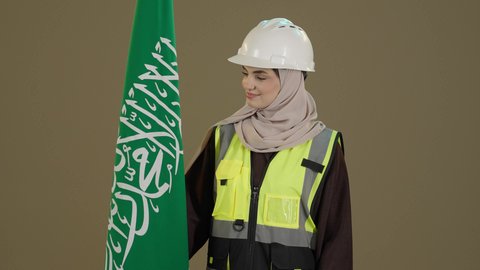 مفهوم حب الوطن، ذكرى تأسيس الدولة السعودية الأولى 22 فبراير، التباهي والتفاخر بامتلاك الهوية السعودية، اليوم الوطني السعودي، بورترية مقرب لمهندسة عربية خليجية سعودية ترتدي الخوذة و سترة الحماية تمسك علم المملكة بتعابير الفخر و السرور، خلفية بني