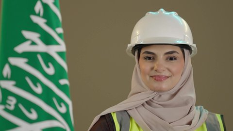 Looking at the camera with gestures of happiness and joy, the anniversary of the establishment of the first Saudi state on February 22, boasting and taking pride in having a Saudi identity, Saudi National Day, a close-up portrait of a Gulf Arab Saudi engineer wearing a helmet, protective vest, and abaya standing next to the Kingdom's flag with expressions of pride and joy, brown background.