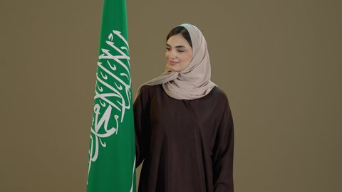 The identity of the Saudi National Day 92, the anniversary of Flag Day on March 11, the celebration of the National Day for the unification of Saudi Arabia by King Abdulaziz on the 8th of Rabi' al-Awwal, a portrait of a smiling Saudi Gulf Arab woman wearing a hijab and an abaya standing in front of the Kingdom's flag with expressions of pride and joy, brown background.