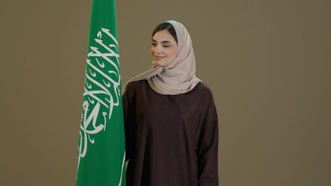 ذكرى يوم العلم 11 مارس، اليوم الوطني السعودي، الاحتفال بالعيد الوطني لتوحيد السعودية على يد الملك عبد العزيز في 8 ربيع الأول، بورترية لسيدة عربية خليجية سعودية مبتسمة محجبة ترتدي العباءة تقف امام علم المملكة تنظرالية بتعابير الفخرو السرور، خلفية بني