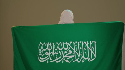 يوم العلم السعودي 11 مارس، الاحتفال بالعيد الوطني لتوحيد السعودية على يد الملك عبد العزيز في 8 ربيع الأول، اليوم الوطني السعودي، سيدة عربية خليجية سعودية مبتسمة محجبة ترتدي العباءة السوداء ترتدي علم المملكة على ظهرها بتعابير الفخرو السرور، خلفية بني