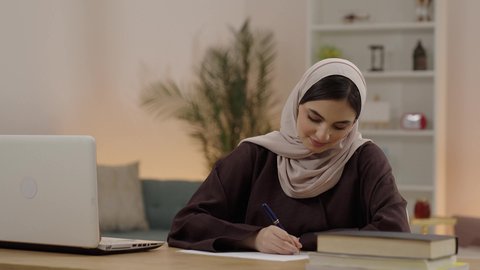 شابة عربية خليجية سعودية محجبة ترتدي العباءة تجلس في مكتبة الجامعة امام الحاسب المحمول تقوم بكتابة التقارير والأبحاث العلمية، التعليم العالي في الكليات السعودية، مراجعة الدروس اليومية، ايماءات التذكر و التفكير فى شيء ما، مفهوم الثقافة والتعلم الذاتي