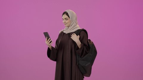 ايماءات السعادة و السرور، عرض الهاتف المحمول بشاشة خضراء, أهمية الدراسة الجامعية, استخدام جهاز تقني حديث متطور, بورتريه لطالبة جامعية عربية سعودية خليجية محجبة ترتدي العباءة  و الحقيبة المدرسية تمسك بيدها الجوال, خلفية ملونه