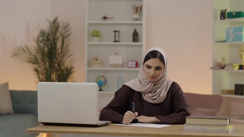 Gestures of remembering and thinking about something, reviewing daily lessons, higher education in Saudi colleges, the concept of culture and self-learning, a young Arab Gulf Saudi woman wearing a hijab and an abaya sitting in the university library in front of a laptop writing reports and scientific research.