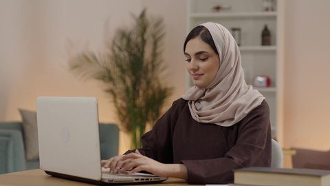 المحاضرات الجامعية الإلكترونية والتعلم عن بعد، مراجعة الدروس اليومية باستخدام الحاسوب المحمول، التعليم العالي في الكليات السعودية، مفهوم الثقافة والتعلم الذاتي، شابة عربية خليجية سعودية محجبة ترتدي العباءة تجلس في مكتبة الجامعة تقوم بكتابة التقارير والأبحاث العلمية