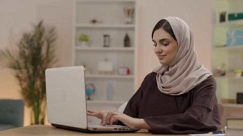 مراجعة الدروس اليومية باستخدام الحاسوب المحمول، المحاضرات الجامعية الإلكترونية والتعلم عن بعد، التعليم العالي في الكليات السعودية، مفهوم الثقافة والتعلم الذاتي، شابة عربية خليجية سعودية محجبة ترتدي العباءة تجلس في مكتبة الجامعة تقوم بكتابة التقارير والأبحاث العلمية