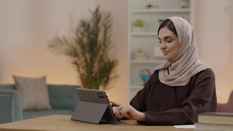 الإستعداد للمحاضرات الجامعية الالكترونية، التعلم عن بعد، اجراء مكالمة فيديو ، طالبة عربية خليجية سعودية محجبة ترتدي العباءة تستخدم جهاز التابلت، التعليم في المملكة العربية السعودية، ايماءات السعادة و السرور