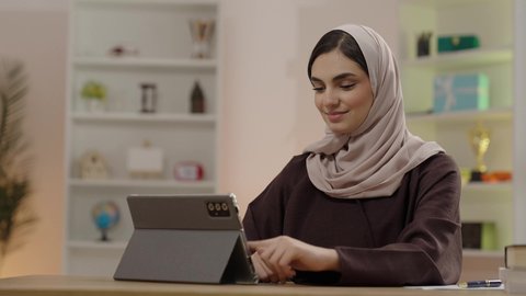 Making a video call, completing electronic assignments, attending online university lectures via a tablet, a veiled Arab Gulf Saudi student wearing an abaya using a tablet, education in the Kingdom of Saudi Arabia, gestures of happiness and joy.