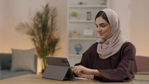 Following online university lectures via a tablet, a Saudi Arabian Gulf Arab student wearing a hijab and an abaya uses a tablet, writing and taking notes, solving electronic assignments, education in the Kingdom of Saudi Arabia, gestures of happiness and joy.