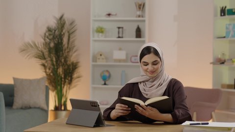 التعليم الجامعي في المملكة العربية السعودية، ايماءات التركيز و الاندماج، مراجعة الدروس اليومية، مفهوم الاجتهاد والتفوق والنجاح, طالبة جامعية عربية خليجية سعودية محجبة ترتدي العباءة تجلس على طاوله خشبية تقوم بمذاكرة الكتاب الجامعي