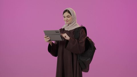 مراجعة الدروس اليومية, التعليم الجامعي في المملكة العربية السعودية, استخدام الأجهزة التقنية الحديثة, بورتريه لطالبة جامعية عربية خليجية سعودية ترتدي العباءة تحمل حقيبة على ظهرها وبيدها الجهاز اللوحي, خلفية ملونه