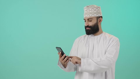 The use of modern devices and advanced technologies, the concept of remote business management, chatting with family and friends, exchanging news via video call, a portrait of an Arab Gulf Omani man wearing a dishdasha and kumma communicating with relatives, expressions of joy and happiness, blue background.