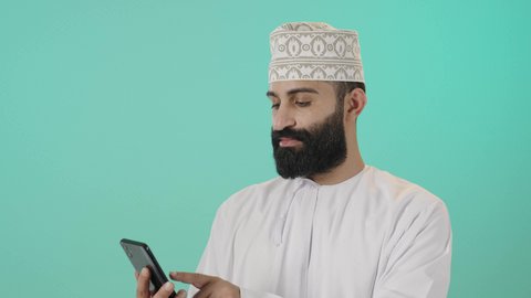 مفهوم ادارة الاعمال عن بعد، استخدام الأجهزة الحديثة والتقنيات المتطورة، الدردشة مع الأهل والأصدقاء، تبادل الاخبار عبرمكالمة فيديو، بورتريه لرجل عربي خليجي عماني يرتدي الدشداشة والكمه يقوم بالتواصل مع الأقارب، تعابير الفرح والسرور، خلفية زرقاء