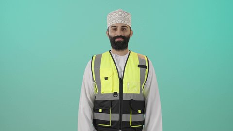 Expressions of self-confidence and seriousness, supervising the progress of the work plan, architectural engineering projects, working in the engineering sector in the Sultanate of Oman, a portrait of an Arab Gulf Omani engineer wearing a dishdasha and kumma and a safety vest standing holding a safety helmet in his hand, a profession and job for youth in the Sultanate of Oman, blue background.