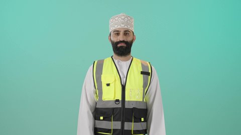 ايماءات الثقة بالنفس والجدية, الاشراف على سير خطة العمل، مشاريع هندسية معمارية, العمل في القطاع الهندسي في سلطنه عمان , بورتريه لمهندس عربي خليجي عماني يرتدي الدشداشة والكمه وسترة الحماية يقف يحمل بيده خوذة الحماية، مهنة ووظيفة للشباب في سلطنة عمان، خلفية زرقاء