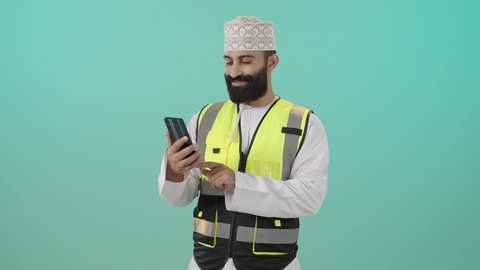 مشاريع هندسية معمارية, الاشراف على سير خطة العمل، ايماءات الثقة بالنفس والجدية, العمل في القطاع الهندسي في سلطنه عمان , بورتريه لمهندس عربي خليجي عماني يرتدي الدشداشة والكمه وسترة الحماية يقف  يقوم بتصفح الهاتف المحمول، مهنة ووظيفة للشباب في سلطنة عمان، خلفية زرقاء