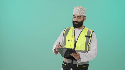 تدوين الملاحظات على ورقة، مفهوم الهندسة والانشاءات المعمارية، بورتريه لمهندس عربي خليجي عماني يرتدي الدشداشة و الكمه وسترة الحماية قاعدة كتابة ينظر إليه بايماءات التركيز والتمعن، مفهوم إدارة المشاريع وتدقيقها، العمل في القطاع الهندسي، خلفية زرقاء